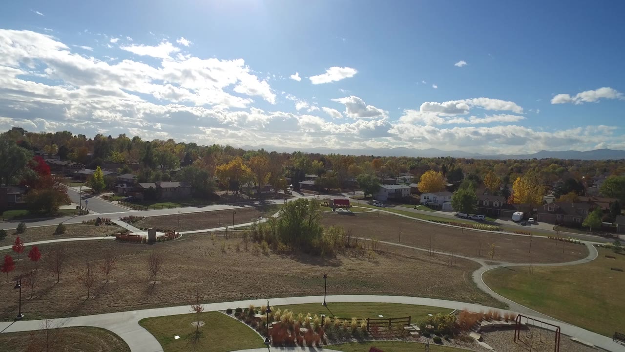 Aerial frame in Hidden Lake, Colorado, with crossing concrete paths in the foreground, open grass, a line of homes, fall-colored trees, distant mountains, and no visible people.