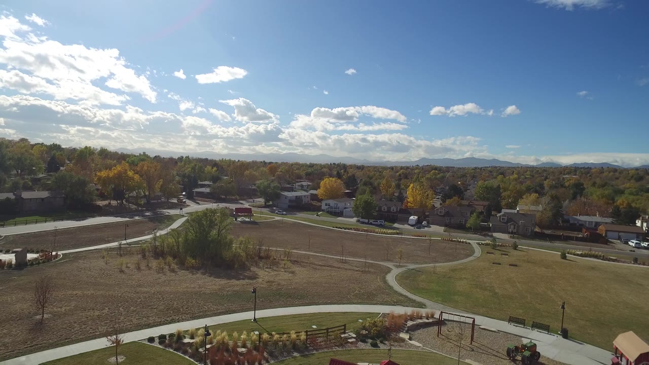 Wide sunny aerial over park grass, paths, roads, homes, and low mountains.