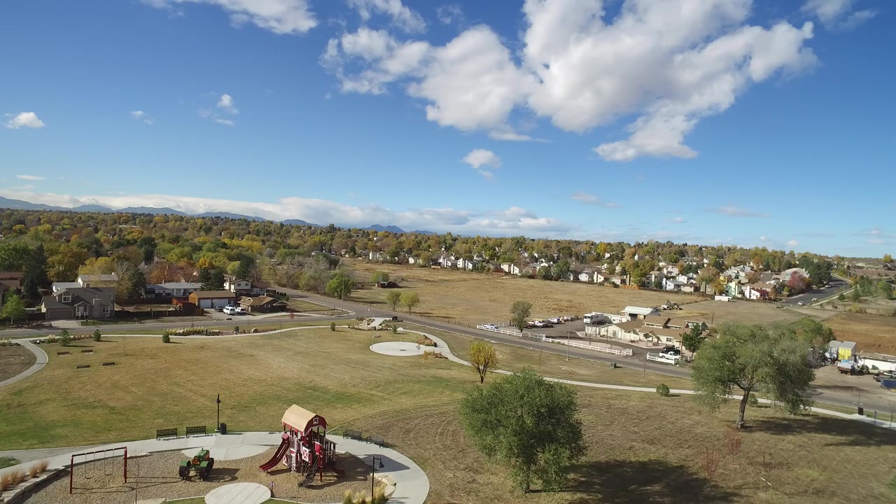 Elevated view over a park playground, open grass, roads, and houses under a blue sky.