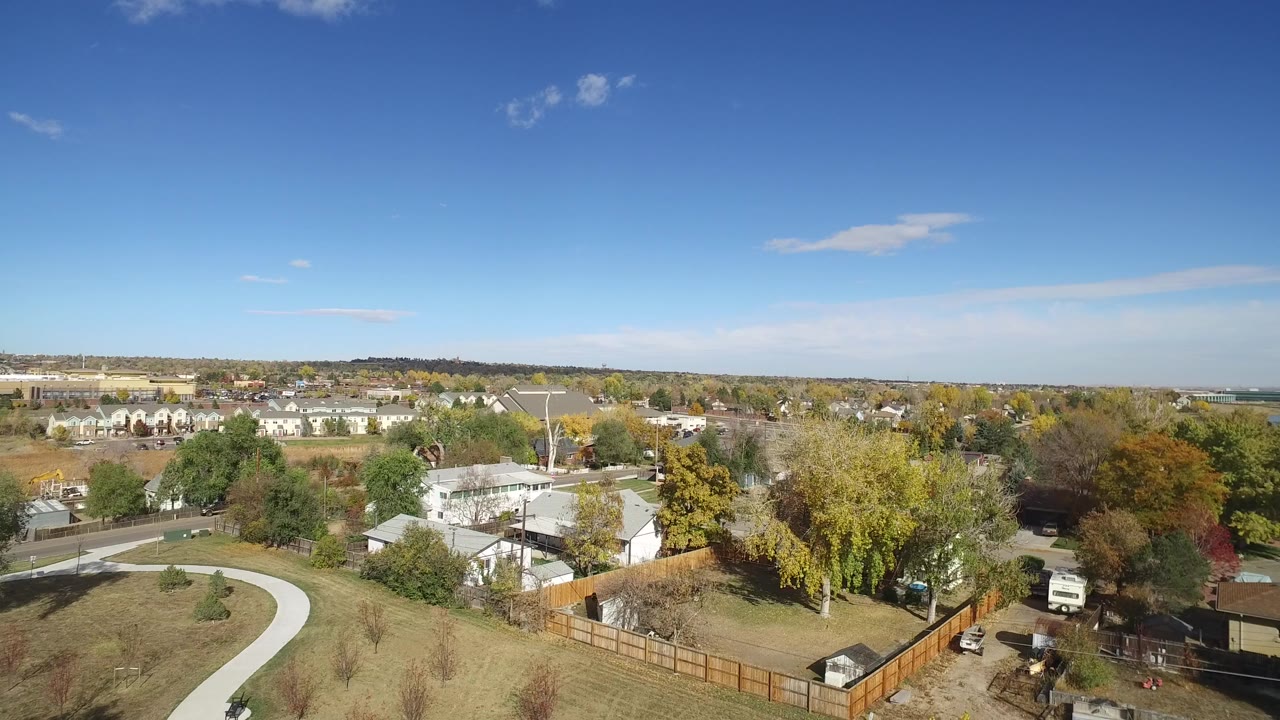 Aerial view of suburban homes, fenced yards, trees, and a distant lake under blue sky.