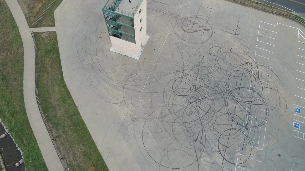 Aerial frame in Hidden Lake, Colorado with dense overlapping round tire marks across the center of the parking lot, bordered by grass, sidewalk, and a small building edge, with no visible people.