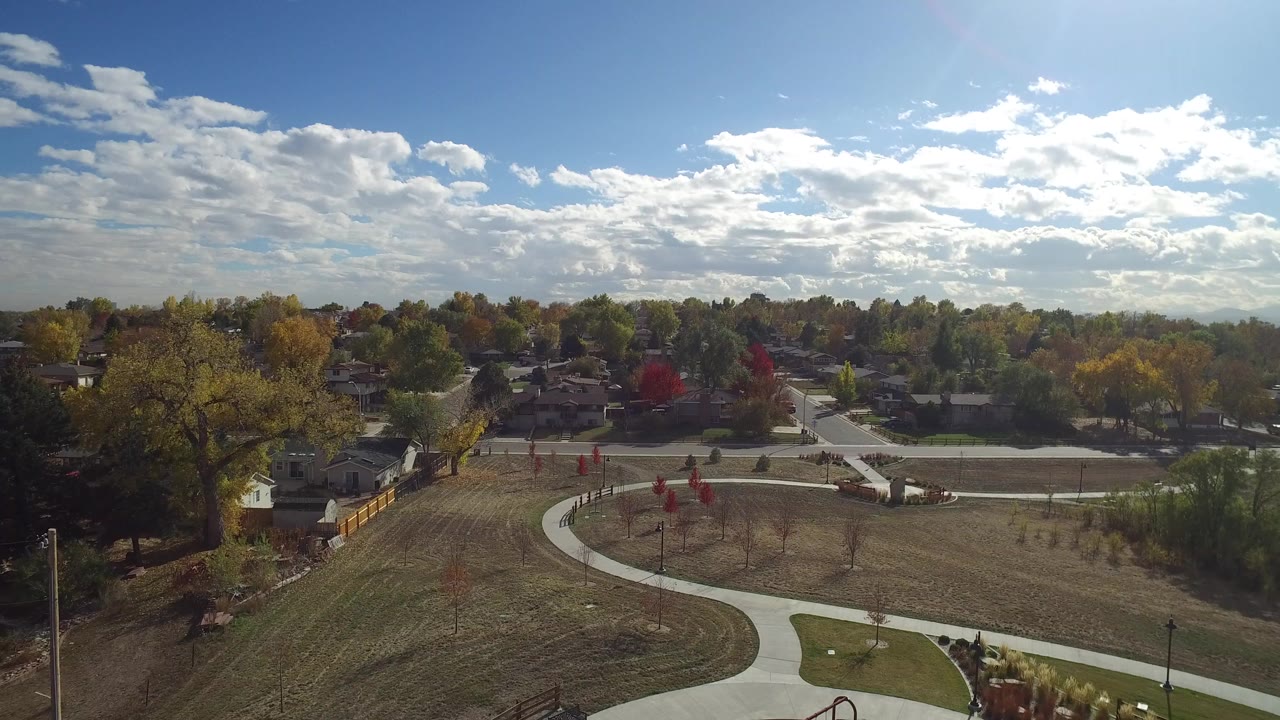 Aerial view of a sunny park with curved paths, grass, trees, houses, and clouds.