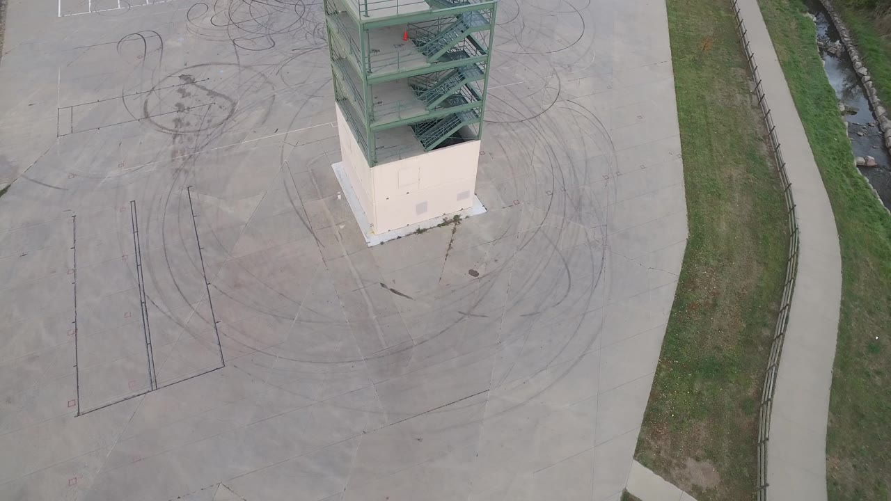 Overhead frame in Hidden Lake, Colorado showing the corner of a green-stair tower above a wide concrete lot with circular tire marks, grass strips, a curved path, and no visible people.