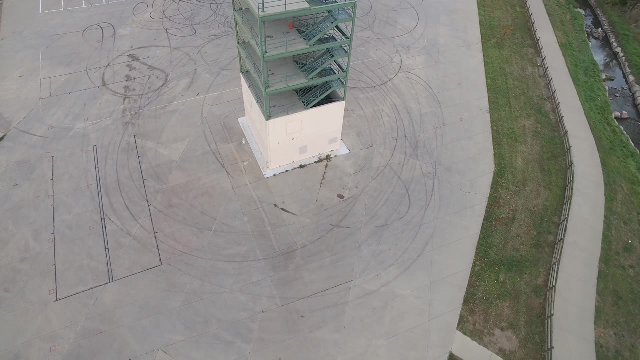 Aerial frame in Hidden Lake, Colorado with the green tower shifted toward the top edge, dark burn-out circles across the concrete, a narrow grass verge, and no visible people.