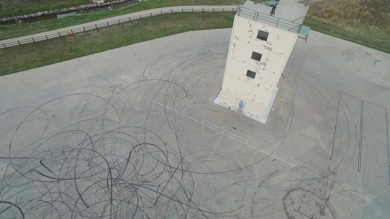 At 0:01, the concrete expanse remains dominant, with looping skid patterns across the lower half, the white structure near the top right, grass and fencing beyond it, and no visible people.