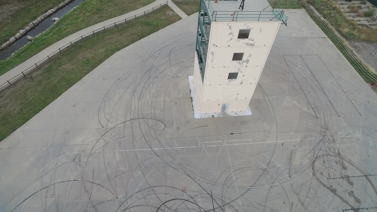 Aerial frame over Hidden Lake, Colorado showing the top of a tall narrow concrete building, pale pavement with tire marks, grass, a canal, and no visible people.