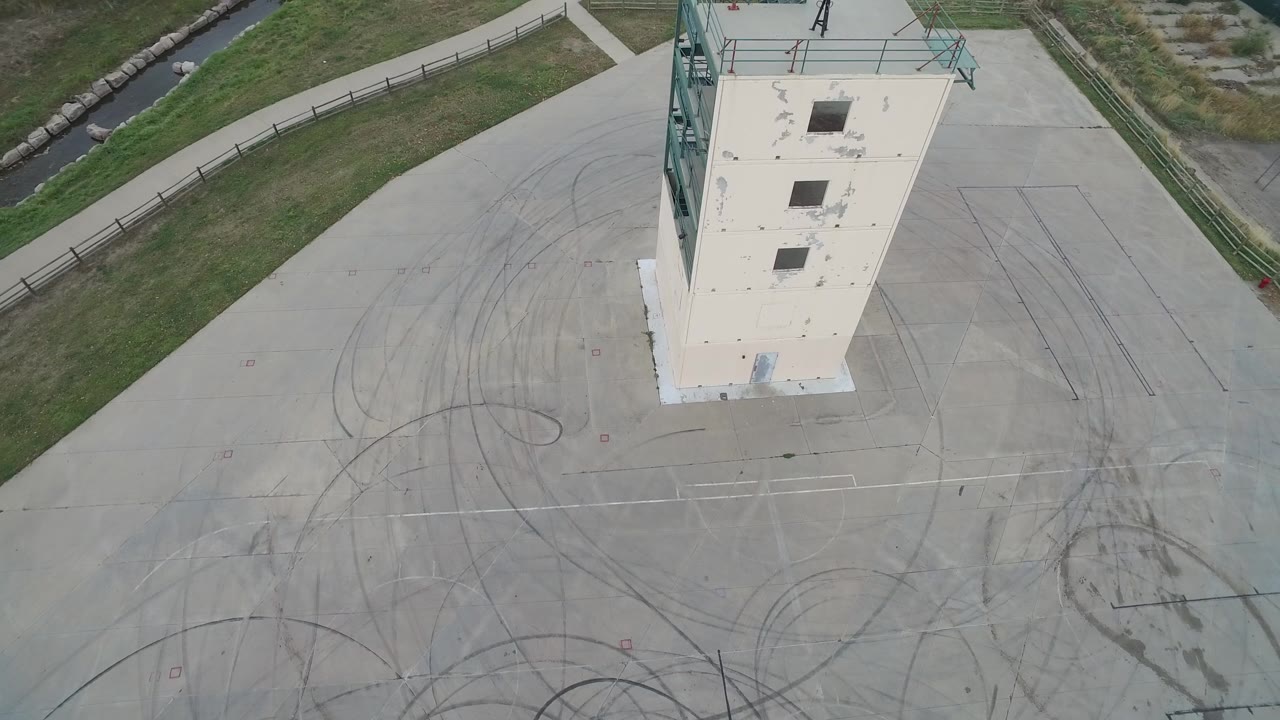 Aerial frame over Hidden Lake, Colorado with the narrow concrete tower shifted slightly in view, rooftop openings, dark tire loops on pavement, grass, canal, and no visible people.