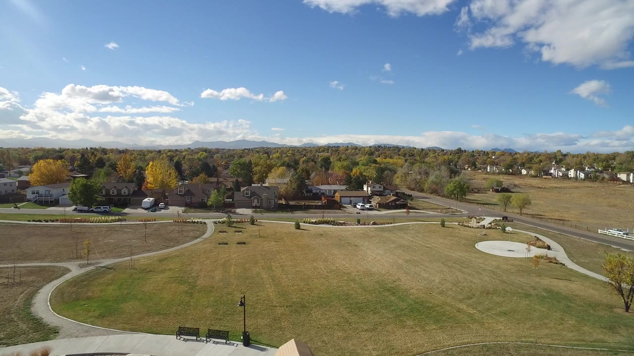 Opening elevated view in Hidden Lake, Colorado, looking across a dry grass park with curving paths, benches, nearby homes, autumn trees, distant hills, and no visible people.