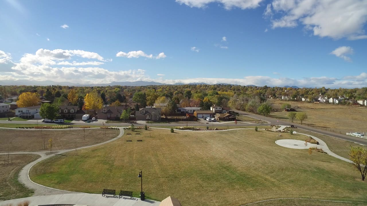Second frame over Hidden Lake, Colorado, holds a wide view of curved sidewalks crossing dry grass, benches near the foreground, homes behind trees, low hills, and no visible people.