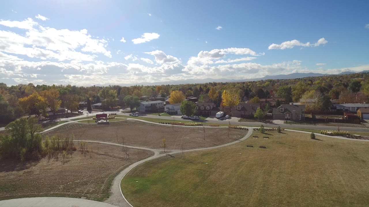 The second frame keeps the Hidden Lake park centered, with a winding concrete path across open grass, autumn trees, houses beyond the road, and no clearly visible people.