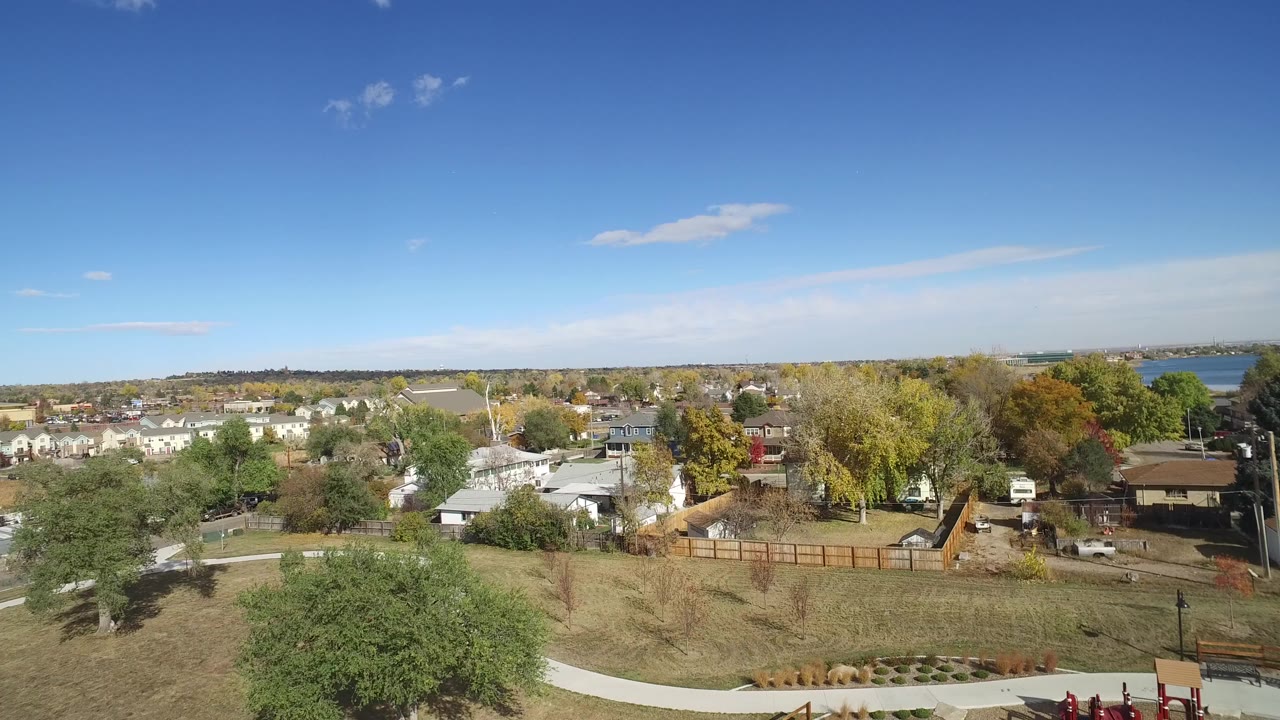 Aerial view over park grass, trees, road, and nearby homes under blue sky.