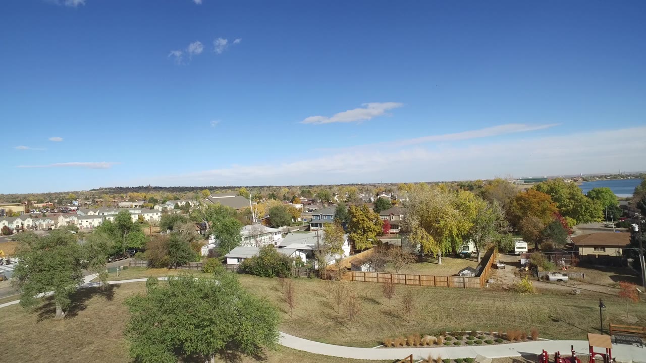 Second aerial frame in Hidden Lake, Colorado, looking across a park lawn and sidewalk toward roadside buildings, trees, fenced yards, and a distant row of homes, with no visible people.