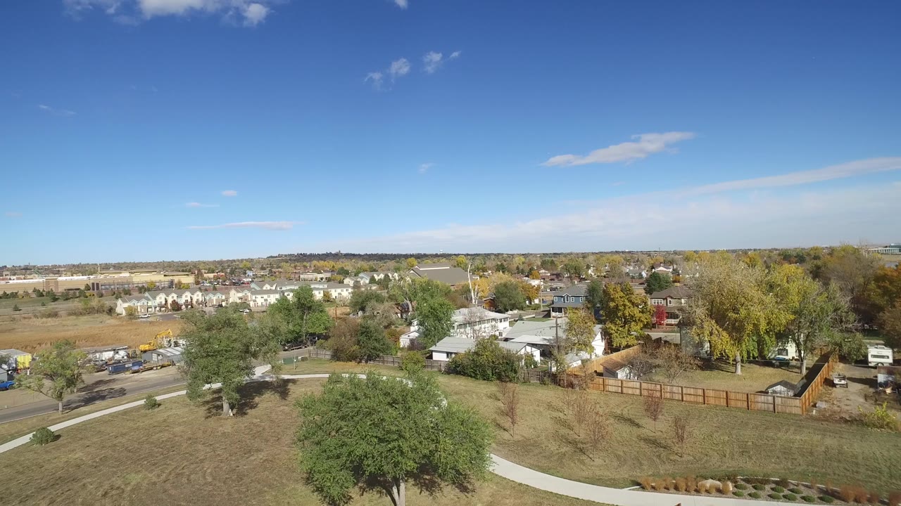 Aerial frame over Hidden Lake, Colorado, with open grass and a sidewalk in front, a road with visible vehicles, trees, small buildings, and homes stretching toward the horizon, with no visible people.