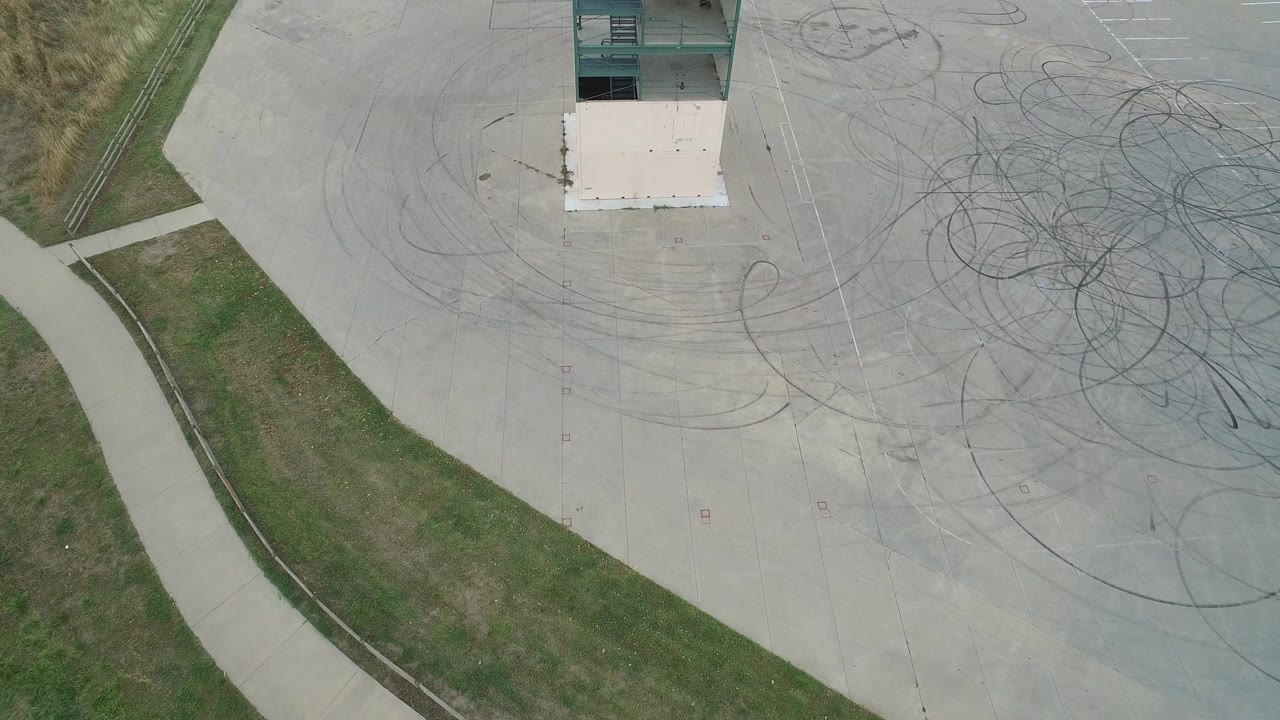 Top-down aerial frame from Hidden Lake, Colorado showing a pale concrete lot, curved tire marks, grass and sidewalk on the left, part of a tower structure at the top, and no visible people.