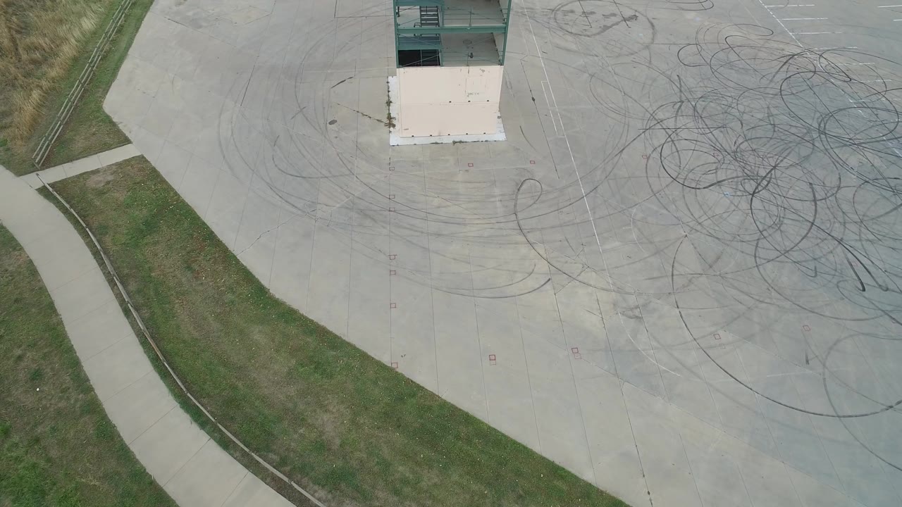 High overhead view of a concrete area with dark circular tire tracks, grass, sidewalk, and a tower section.