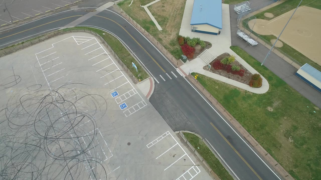 Aerial view of a parking lot, curved road, grass, and part of a baseball field