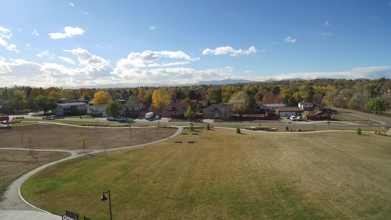 Aerial view of a grassy field with curved paths, homes, trees, and distant hills.