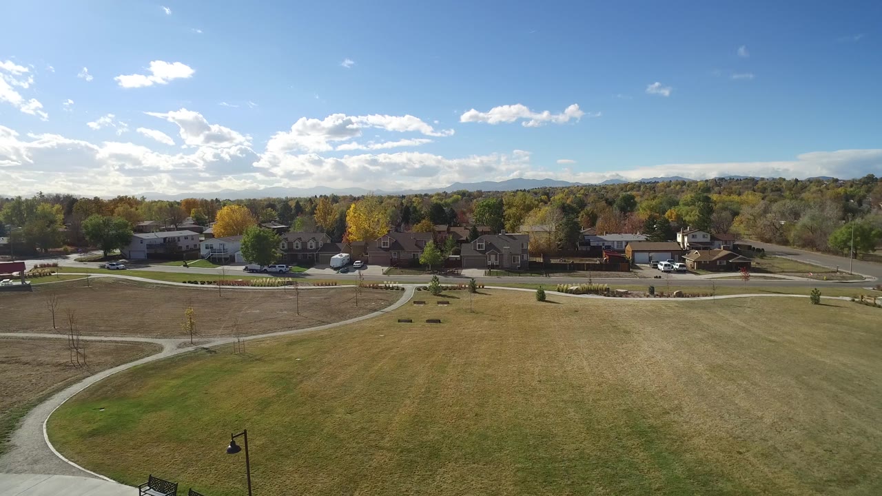 Wide aerial frame in Hidden Lake, Colorado with curved walking paths across grass, a road and homes beyond, yellow and green trees, distant hills, and no visible people.