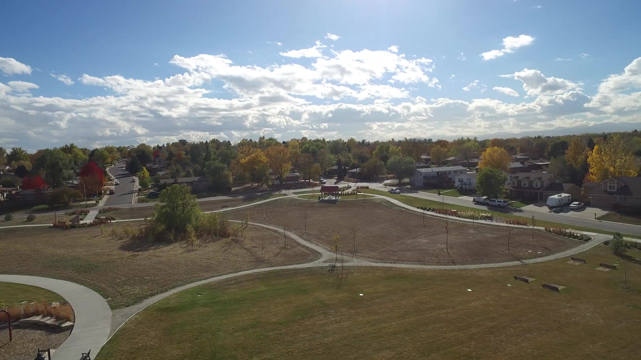 High aerial view of a suburban park, curved paths, open grass, trees, and homes under a bright sky.