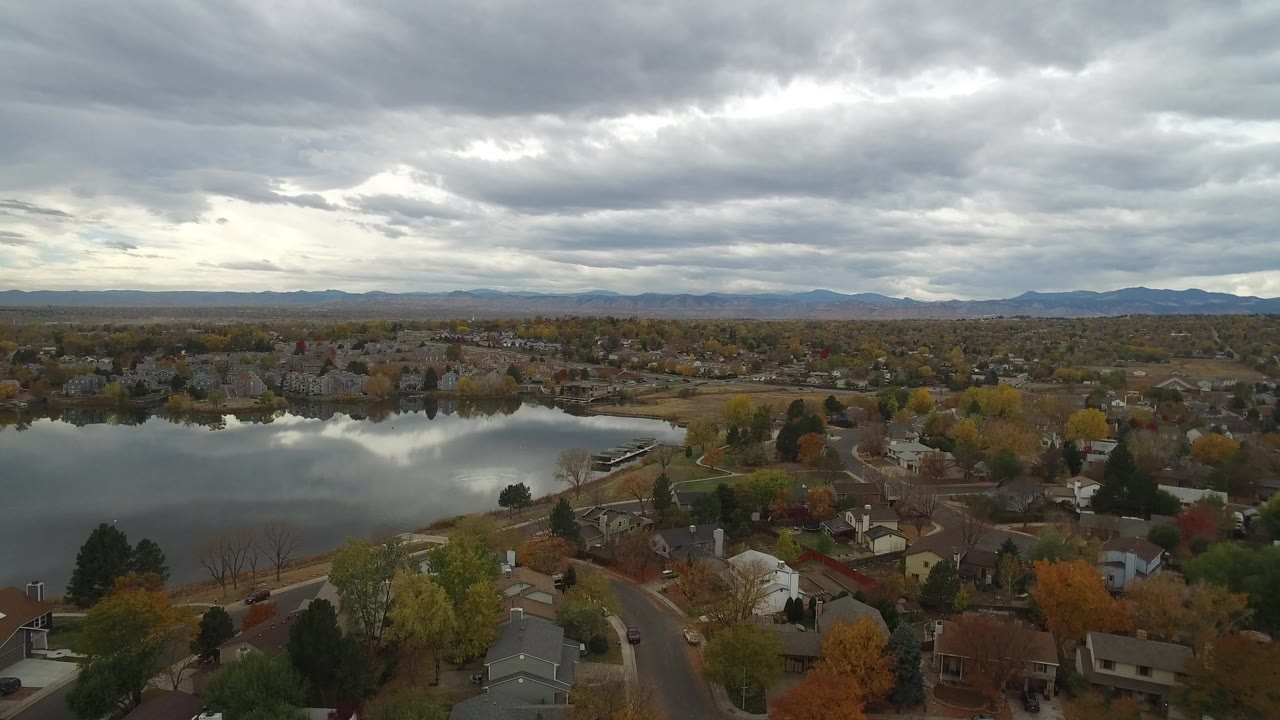 Aerial view of a lake, houses, autumn trees, and distant mountains under gray clouds.