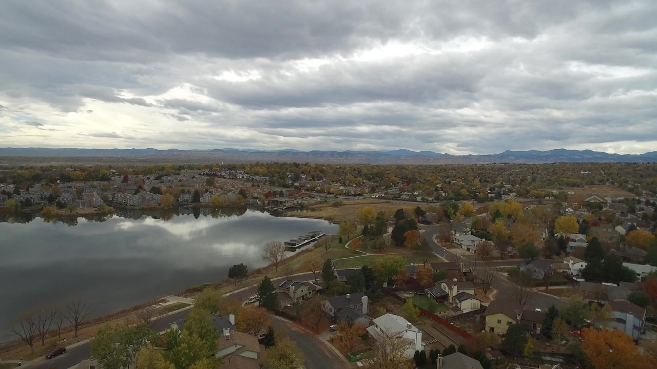 Aerial frame over Hidden Lake, Colorado showing a reflective lake at left, curved streets, detached homes, autumn trees, distant foothills, and no visible people.