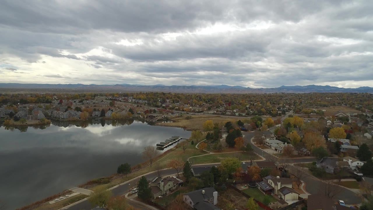 Opening aerial frame over Hidden Lake, Colorado, with calm water at left, residential streets and homes below, distant mountains on the horizon, and no visible people.
