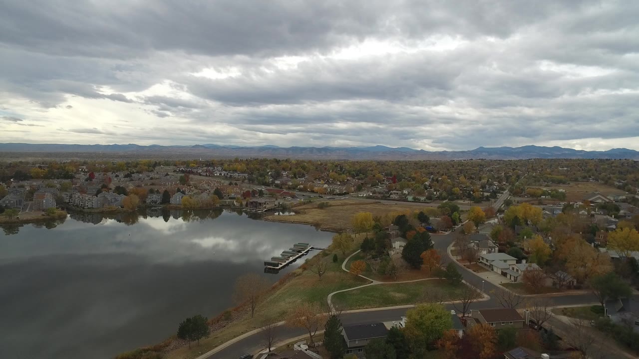 Opening aerial frame over Hidden Lake, Colorado, with calm water at left, homes and autumn trees at right, distant mountains on the horizon, and no visible people.