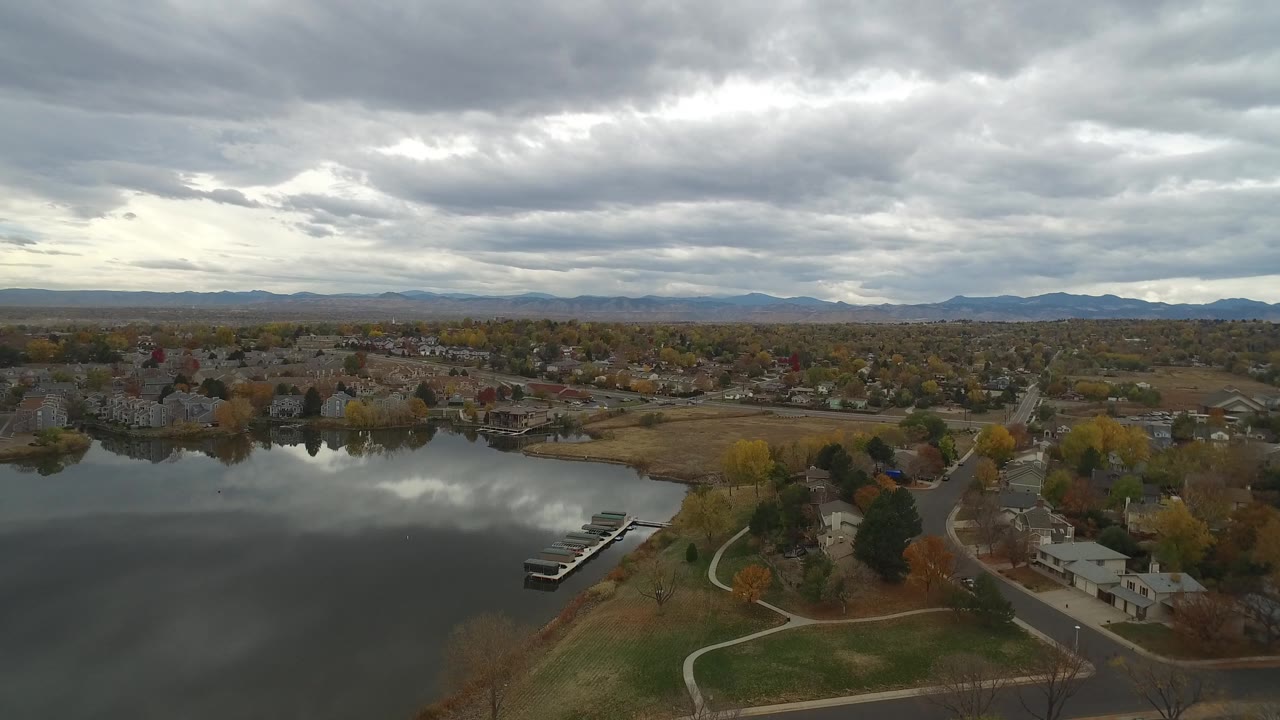 Drone-height view across Hidden Lake, Colorado, with reflective water in the foreground, small docks near shore, residential blocks beyond, and no visible people.
