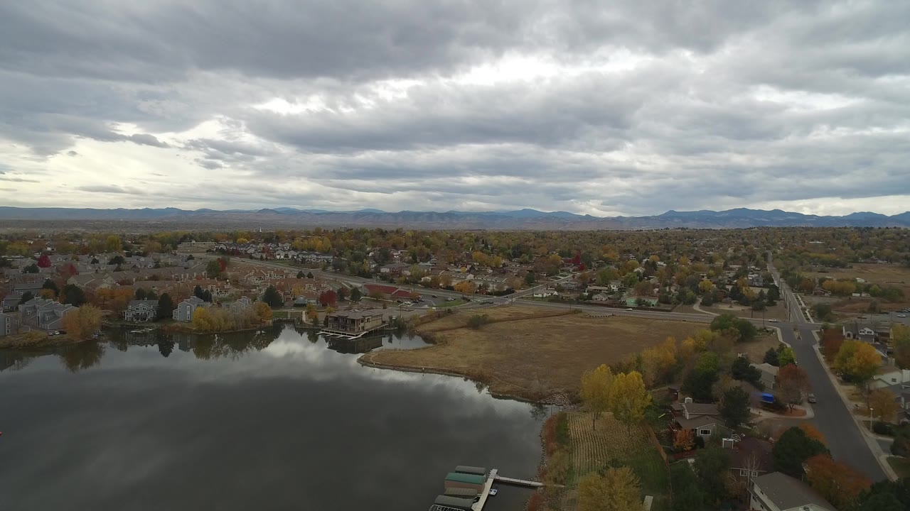 Aerial view over a lake, homes, fields, trees, and distant mountains under gray clouds.