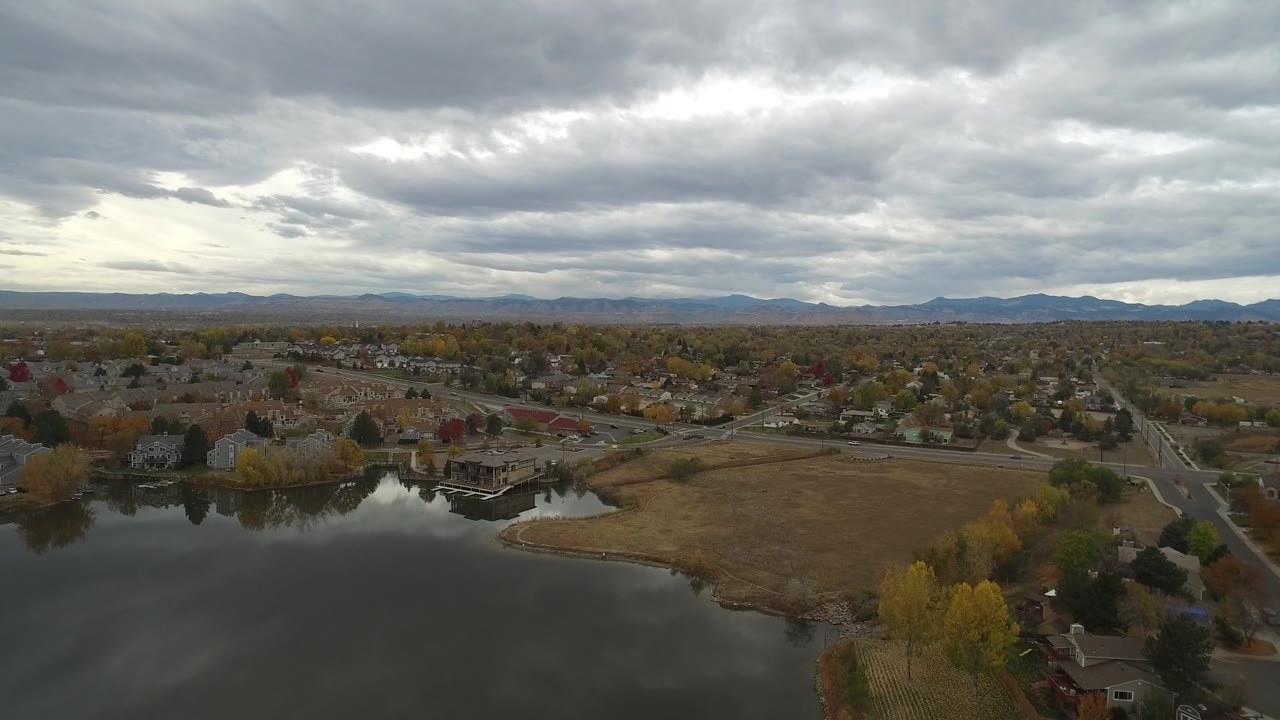 Second aerial frame over Hidden Lake, Colorado, showing the lake edge, clustered homes, a road junction, autumn trees, low mountains, and no visible people.