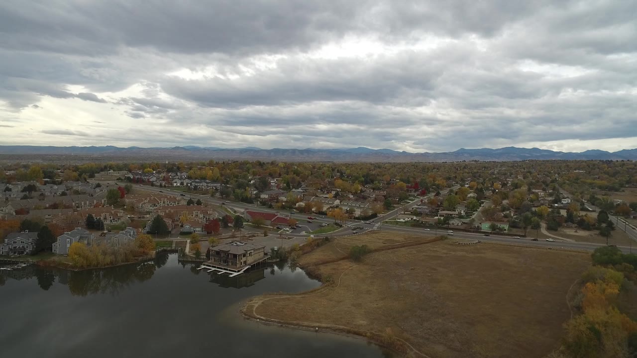Aerial view over Hidden Lake, Colorado with dark water at lower left, brown open grass, nearby houses, roads, autumn trees, distant mountains, and no visible people.