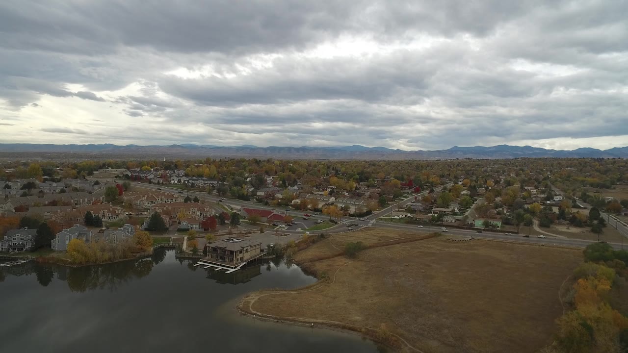 Wide aerial frame from Hidden Lake, Colorado showing a curved shoreline, residential streets, houses, fall-colored trees, low mountains on the horizon, heavy gray sky, and no visible people.