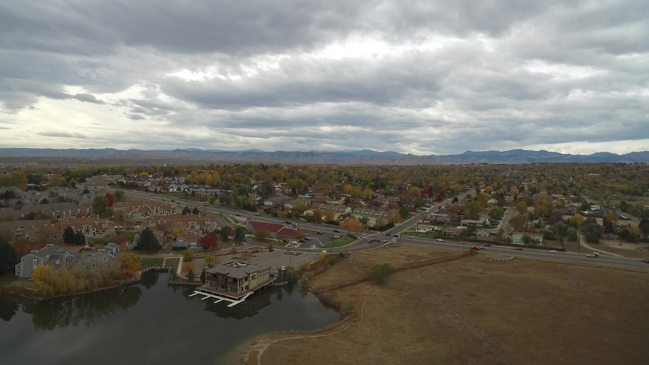 Aerial view over a lake edge, homes, roads, fall trees, and cloudy mountains