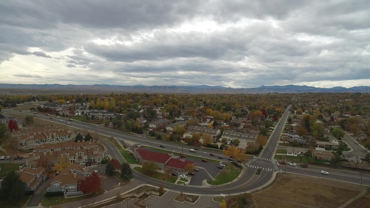 Aerial view of a suburban intersection, houses, trees, and distant mountains under gray clouds.