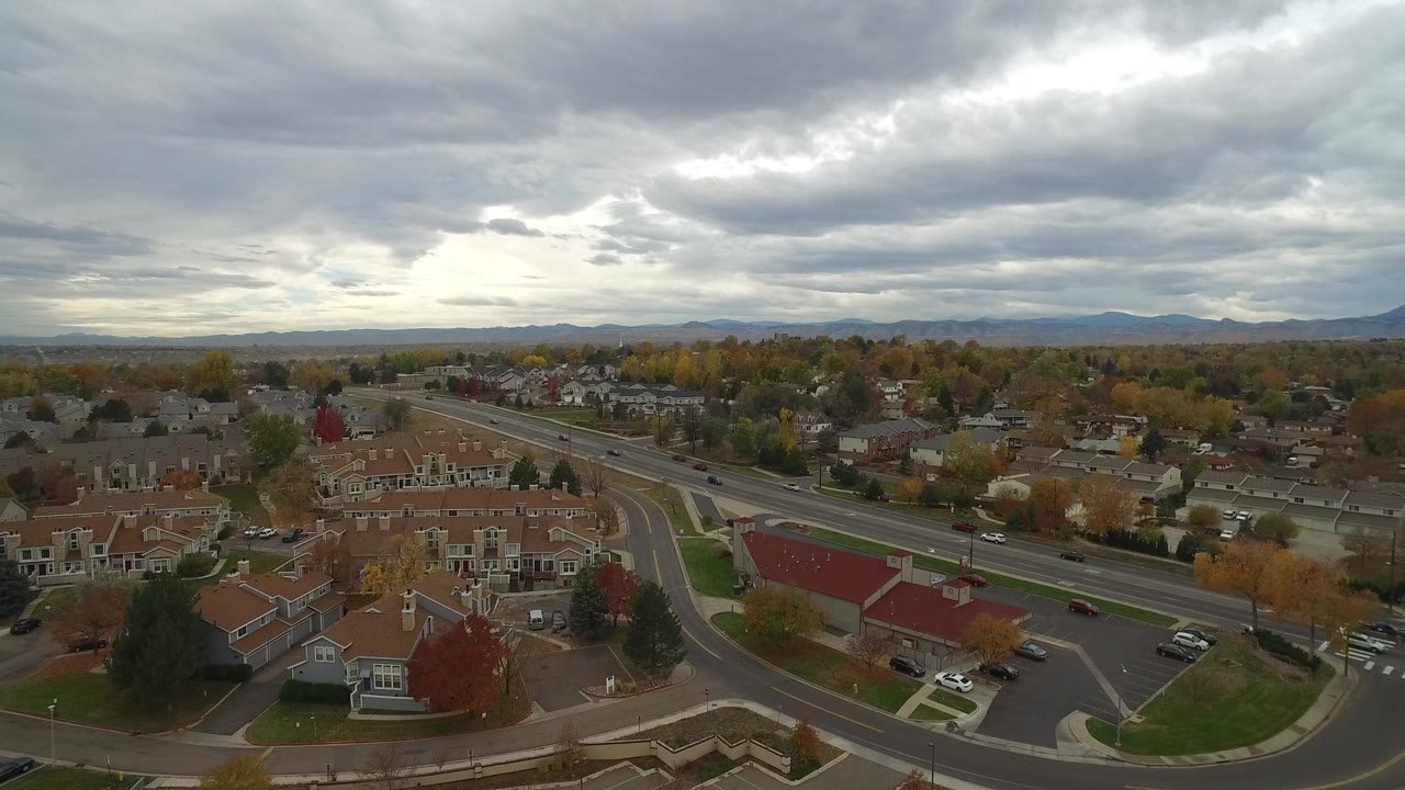 Opening aerial frame over Hidden Lake, Colorado with rows of townhomes, curving roads, autumn trees, distant hills, a gray cloud deck, and no visible people.