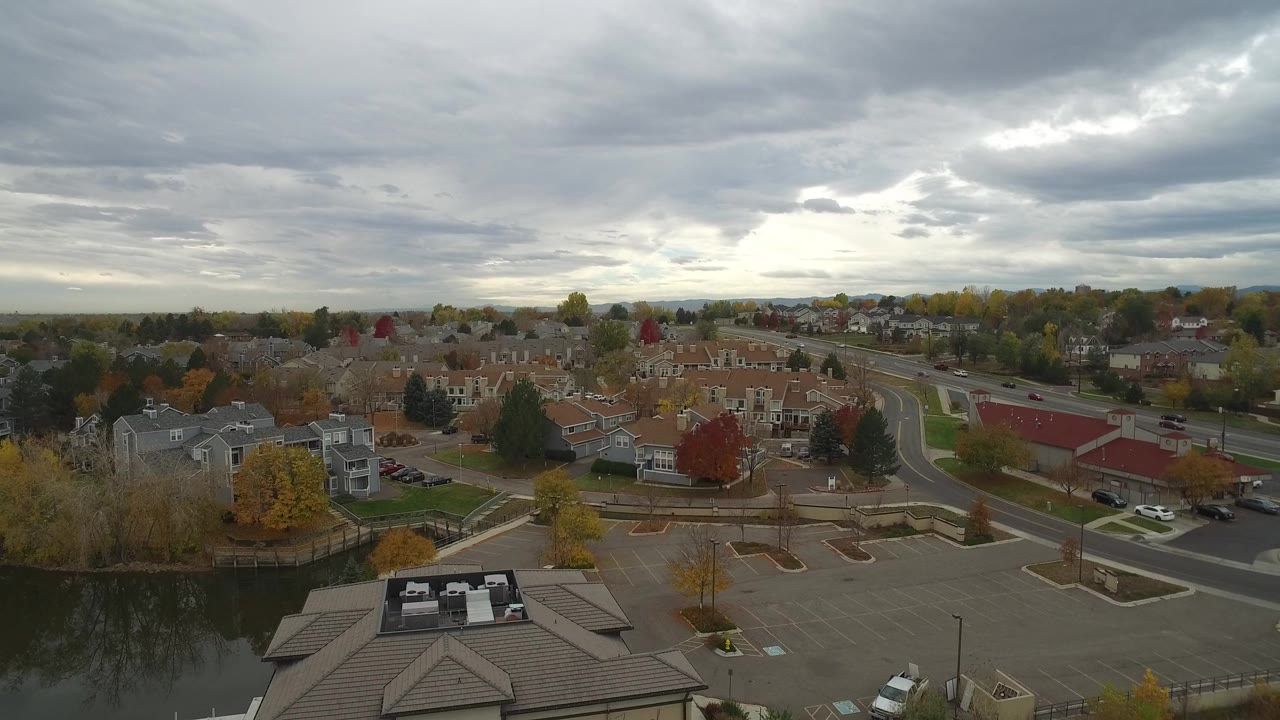 Wide aerial view of a lakeside building, roads, homes, water, and cloudy sky.