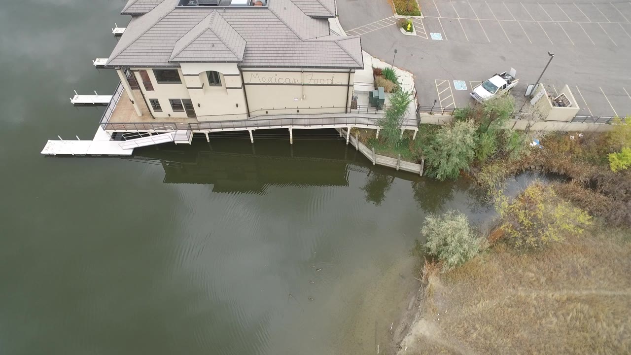 Aerial view of a lakeside building, dock, water, brush, parking lot, and white car.