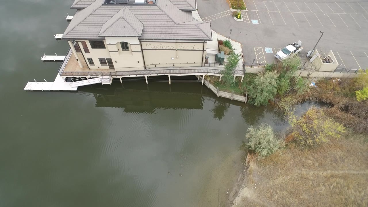 A second aerial frame over Hidden Lake, Colorado keeps the beige building near the top edge while water, reeds, marked parking spaces, and a white car remain visible with no visible people.