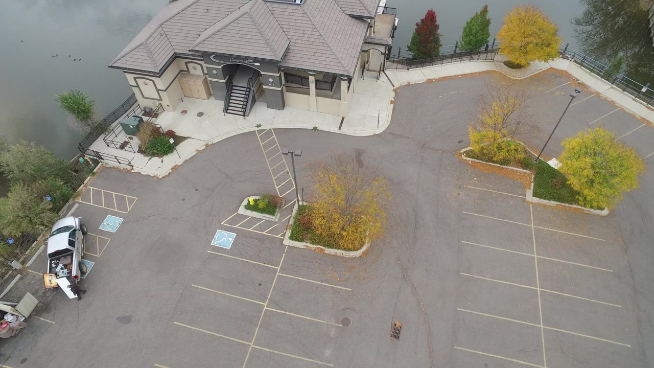 High aerial frame over Hidden Lake, Colorado showing a large lakeside building, empty parking rows, autumn trees, a gray lake edge, and a small person near a white vehicle.