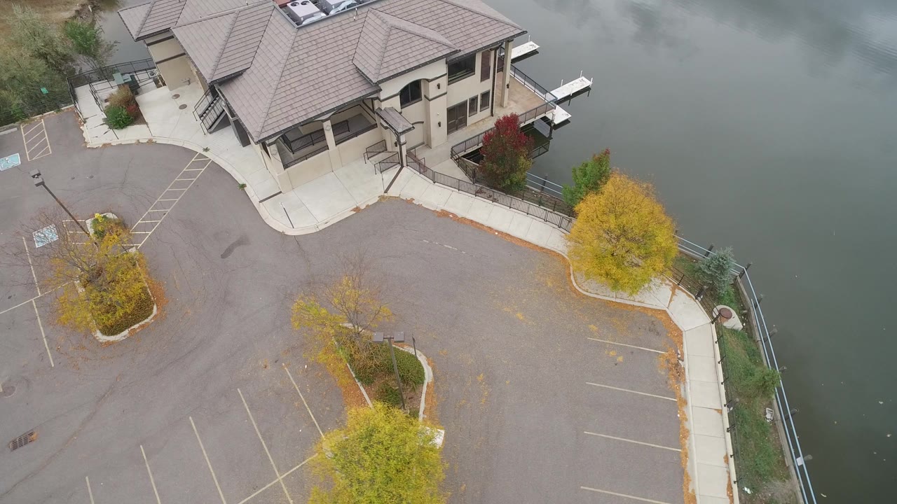 Overhead view of a waterfront building, empty parking lot, trees, and water.