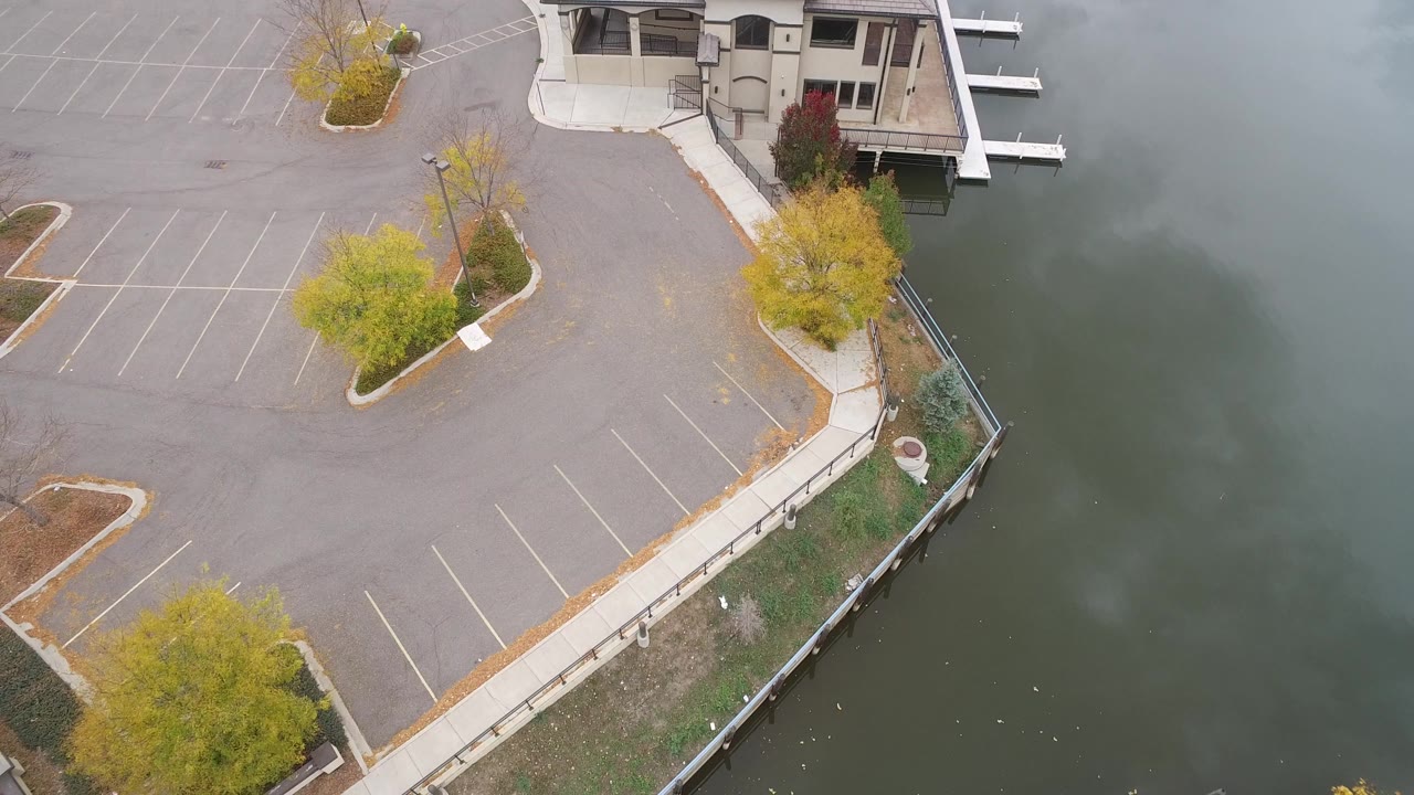 Aerial frame over Hidden Lake, Colorado showing marked parking spaces, landscaped trees, a shoreline walkway, part of a large waterfront building, green-gray lake water, and no visible people.