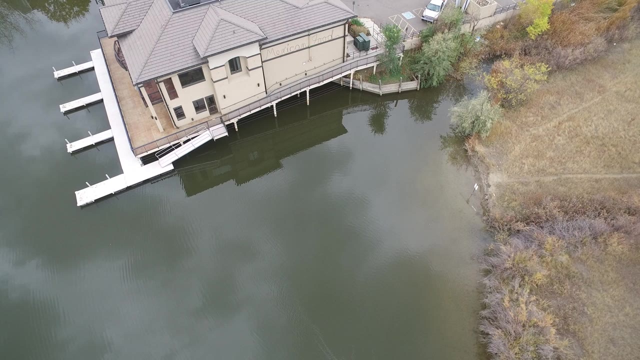 Overhead frame in Hidden Lake, Colorado showing a beige waterfront house at the top edge, white dock sections, green-gray lake water, dry grass shoreline, and no visible people.