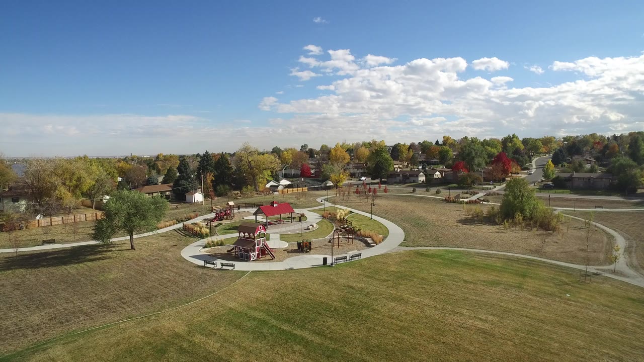 Aerial view of a suburban park, playground, lawns, trees, roads, and homes under a blue sky.