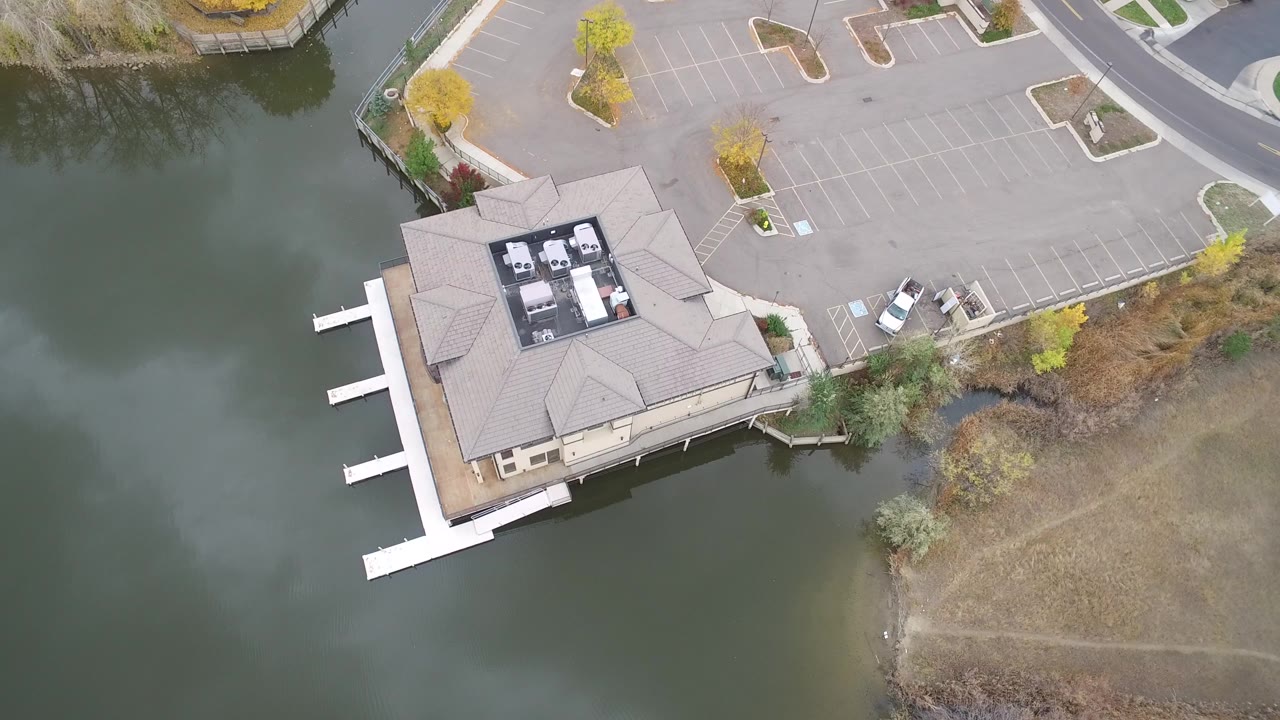 First frame of Hidden Lake, Colorado aerial footage showing a beige-roofed waterfront building, three white docks, green lake water, empty parking spaces, fall trees, and no visible people.