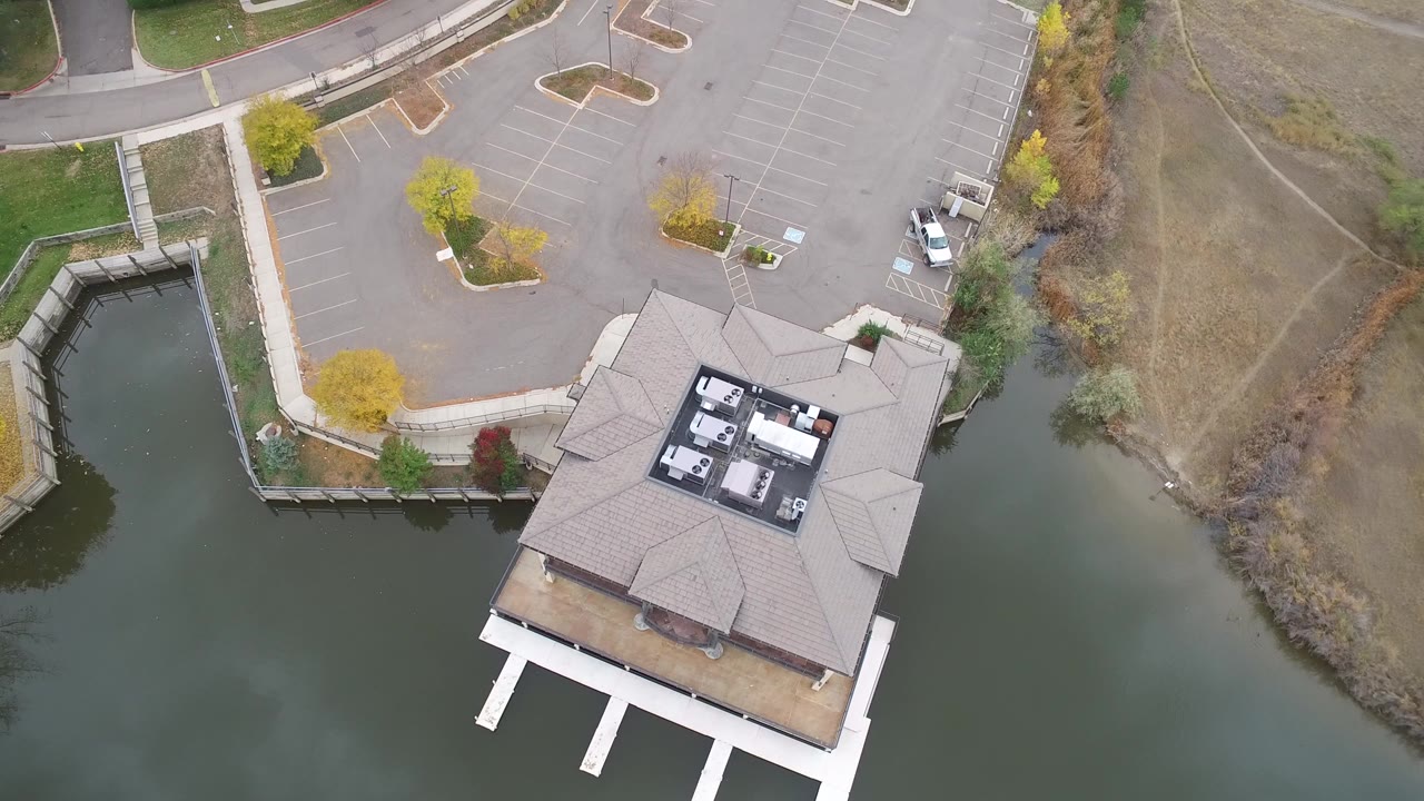 Overhead frame from Hidden Lake, Colorado showing a roofed building over green-blue water, a paved parking lot, sidewalks, autumn trees, and no visible people.