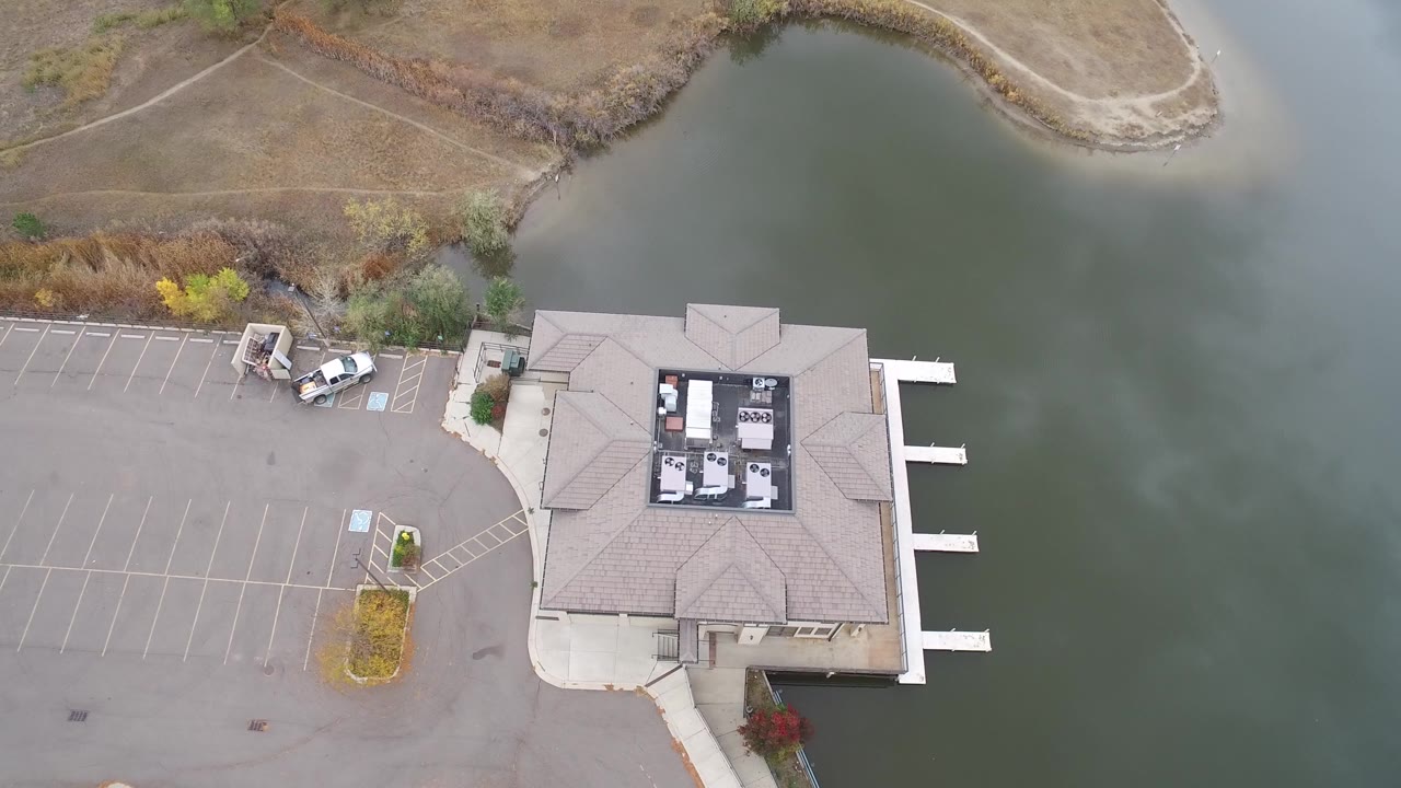 High aerial frame over Hidden Lake, Colorado, showing a tan-roofed building beside green lake water, a mostly empty parking lot, a few parked vehicles, docks, shoreline brush, and no visible people.