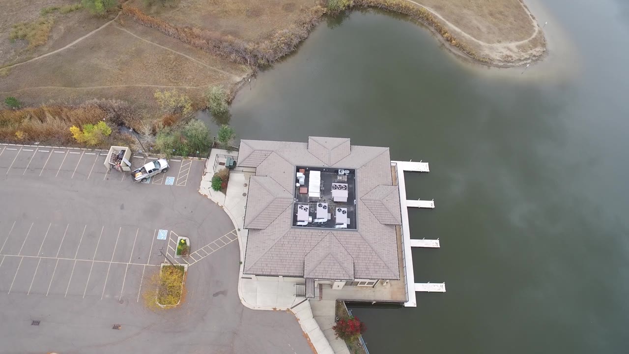 Hidden Lake, Colorado aerial frame with more of the paved lot visible at left, the lakeside building at lower right, muted green water behind it, parked vehicles near the shore, and no visible people.