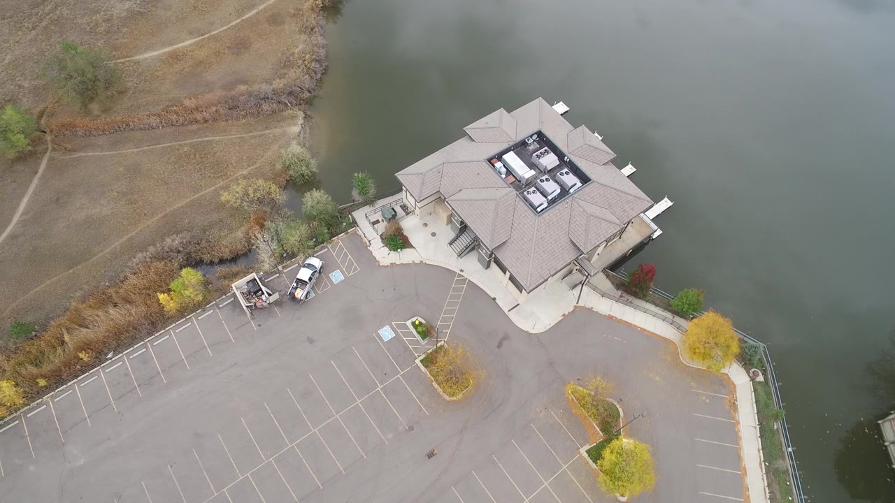 Overhead frame from Hidden Lake, Colorado, showing a gray-roofed lakeside building, empty marked parking stalls, shoreline grass, and green-gray water with no visible people.