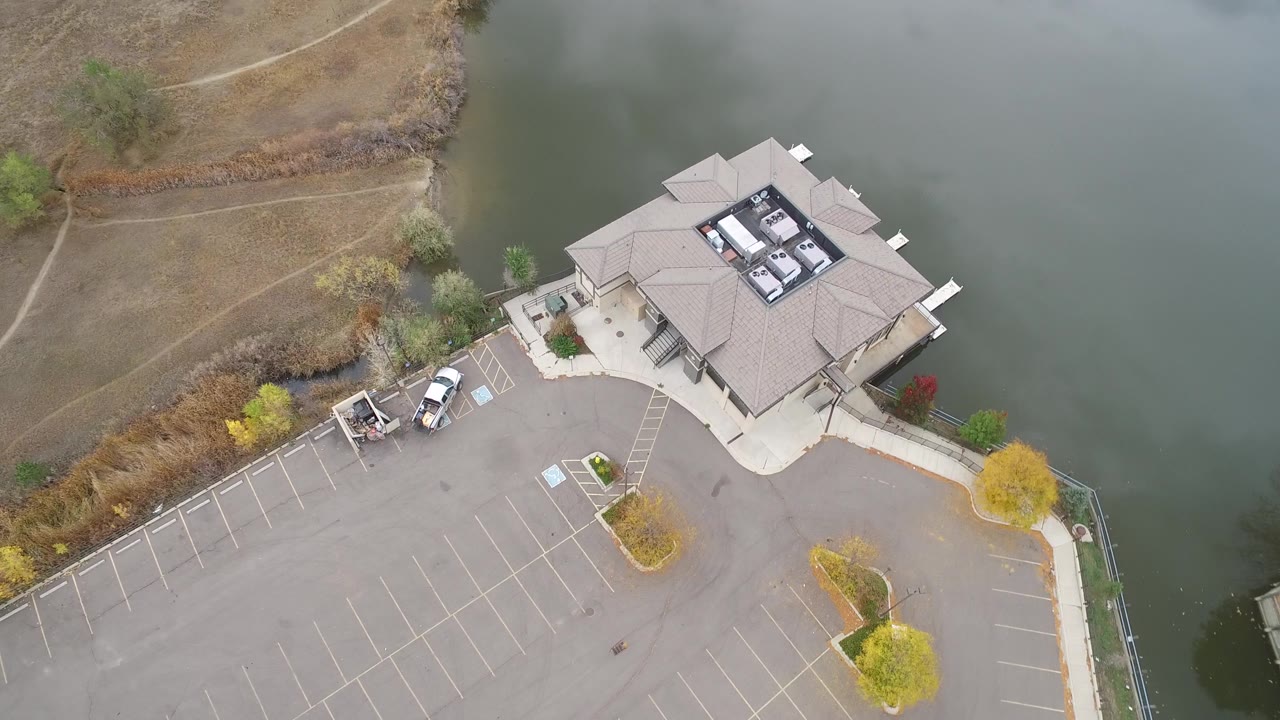 Aerial frame from Hidden Lake, Colorado, with the building slightly right of center, broad empty asphalt below, shoreline vegetation to the left, and no visible people.