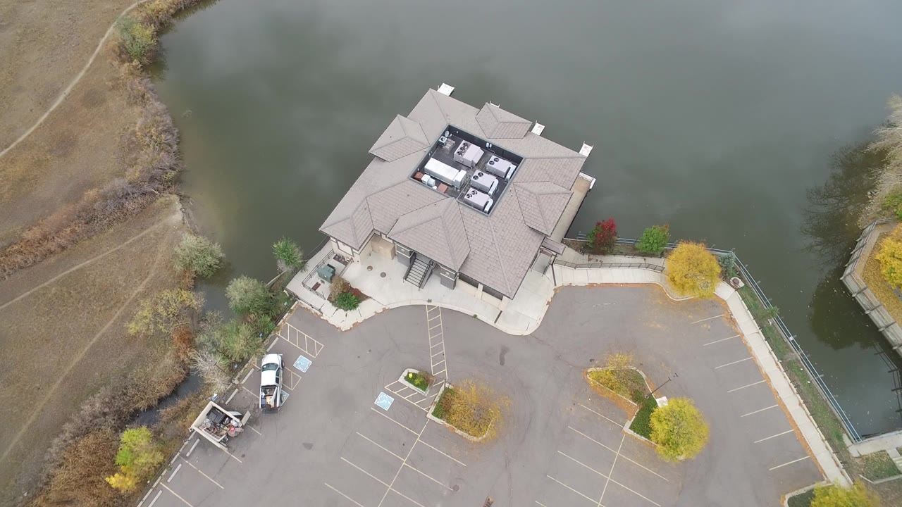 Top-down aerial frame of a light-roofed building beside green-gray water and a mostly empty parking lot in Hidden Lake, Colorado, with parked vehicles and fall-colored trees visible.